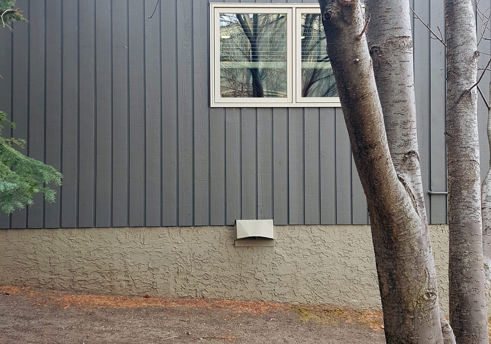 Exterior Wall Of Home With Cold Air Duct. Gray house wall with tree in foreground.